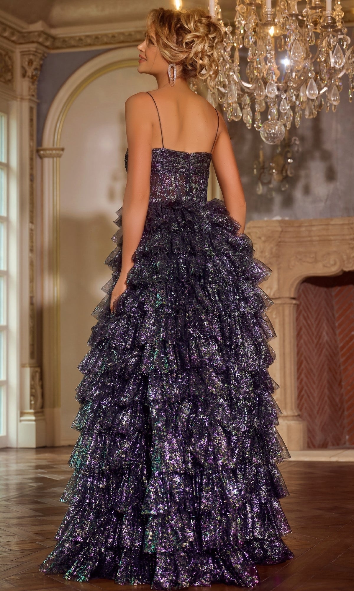 A woman in the Jovani 39305 Ruffled Glitter-Tulle Prom Ball Gown stands with her back to the camera in an elegant, chandelier-lit room.